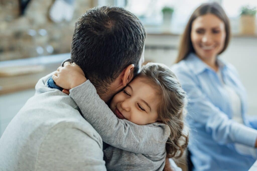 Photo d'une petite fille faisant un câlin à son père avec sa mère en arrière plan ce qui permet de créer des liens d’attachement solides